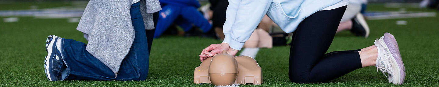 Two individuals kneel on artificial turf, one demonstrating CPR techniques on a training mannequin while the other observes closely.